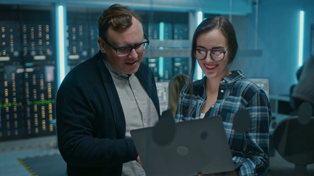 男IT工程师和女IT程序员交谈，拿着笔记本电脑。在后台技术部门，有服务器机架和人员工作。概念:人工智能、机器学习视频素材