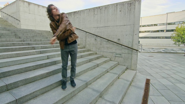 愉快和快乐的长头发的年轻人积极跳舞，而走下楼梯。他穿着一件棕色皮夹克。场景拍摄于商业中心旁边的城市混凝土公园。阳光明媚的。视频素材