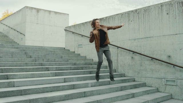 愉快和快乐的长头发的年轻人积极跳舞，而走下楼梯。他穿着一件棕色皮夹克。场景拍摄于商业中心旁边的城市混凝土公园。阳光明媚的。视频素材
