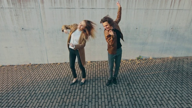 快乐的女孩和快乐的长头发的年轻人正在城市混凝土墙旁边的街道上积极地跳舞。他们穿着棕色的皮夹克和外套。这是在一个阳光明媚的日子里拍摄的。视频素材