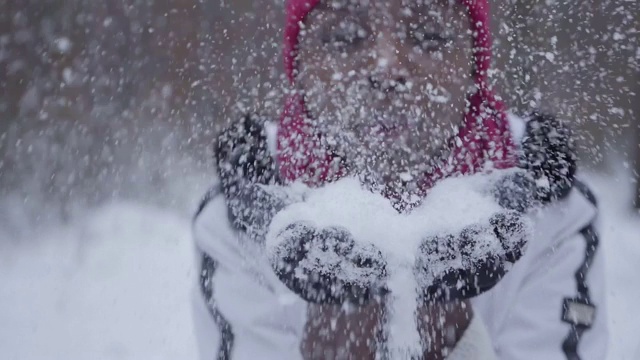照片中的非洲裔美国女孩穿着暖和，戴着无檐小帽和白色夹克，把手中的雪花吹进了站在冬日森林里的相机里。视频下载