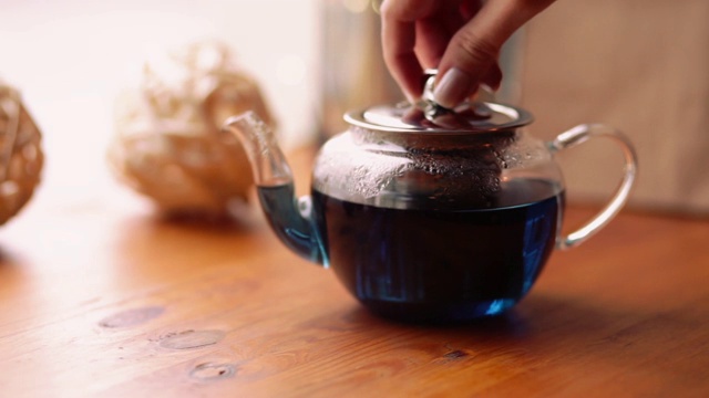 女人打开茶壶盖，盖上热的蓝色花泰国茶。蓝蝴蝶豌豆茶，从花阴蒂里亚在咖啡馆的桌上。视频素材