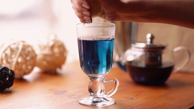 一位女士将一个柠檬挤进了一杯泰国蓝花茶蓝蝴蝶豌豆茶中。蓝茶对柠檬汁的反应。蓝色的茶变成紫色。慢动作视频素材