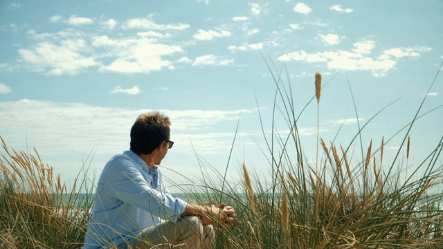 男人坐在沙滩上的沙丘上。视频下载
