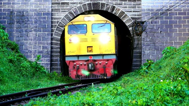 慢动作火车开着大灯，风吹着从旧隧道驶出。古铁路隧道前的山脚下绿草如茵。视频下载