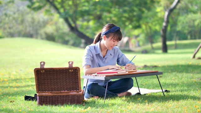 年轻美丽的亚洲女人写在笔记本上，伸展自己在自然或公园和阳光。清新愉快的放松。慢动作视频下载