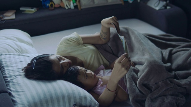 年轻的亚洲母亲和她的儿子坐在床上看书。家庭阅读时间。视频素材