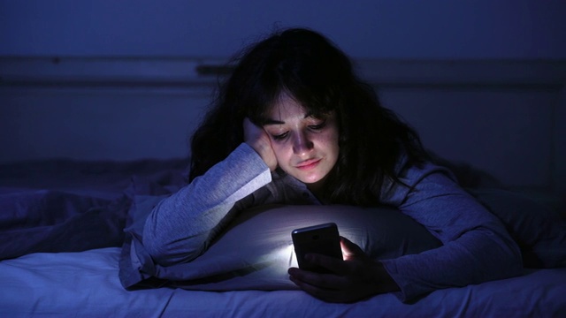 一个沉迷的年轻女人聊天和上网用她的智能手机在深夜昏昏欲睡，无聊和疲惫的4K宽视频。戏剧性的暗光。在互联网、移动成瘾和失眠症的概念。视频下载