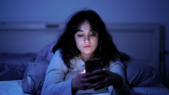 一个沉迷的年轻女人聊天和上网用她的智能手机在深夜昏昏欲睡，无聊和疲惫的4K宽视频。戏剧性的暗光。在互联网、移动成瘾和失眠症的概念。视频购买