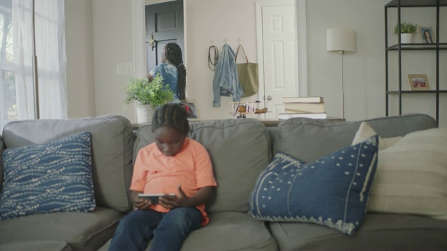 一位非裔美国妇女在沙发上玩智能手机游戏的儿子面前接受快递和摇盒子。视频素材