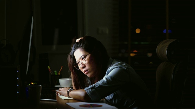 迷人的亚洲女人工作到很晚，思考与严肃的行动在桌子前的电脑显示器桌面在黑暗的工作场所，工作到很晚和努力的概念视频下载