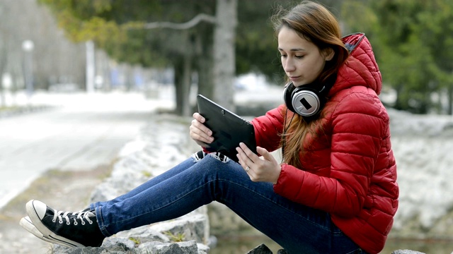 一个十几岁的女孩穿着红色夹克，头戴红色头发，坐在城市春天公园一个空喷泉旁的石头上玩着平板电脑。肖像。关闭了。29.97帧/秒。视频素材