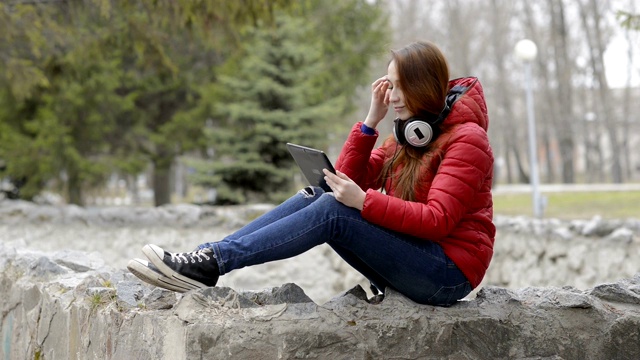 美丽的女孩休息和玩平板电脑，坐在他的头戴耳机在城市春天公园。她有红色的头发和一件红色的夹克。肖像。侧视图。29.97帧/秒。视频素材