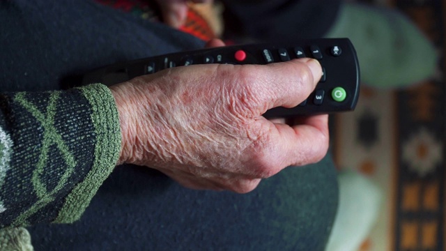 一个老年人看电视，娱乐，一个奶奶的手满皱纹使用遥控器的特写。视频下载