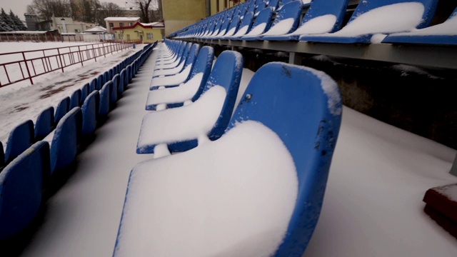 在冬季，体育场看台上一排排被雪覆盖的蓝色塑料座位视频素材