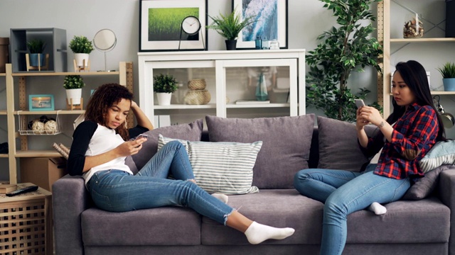 穿着休闲的女性朋友坐在公寓的沙发上玩智能手机。现代科技，年轻的生活方式和时尚的小玩意概念。视频素材