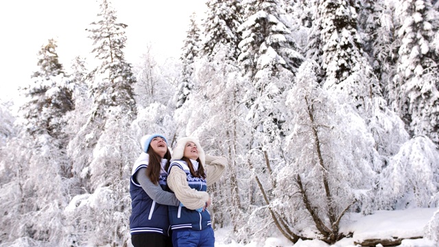 白雪覆盖的森林里的一条河边，两个快乐的女孩拥抱在一起笑着。情人女孩享受散步视频下载