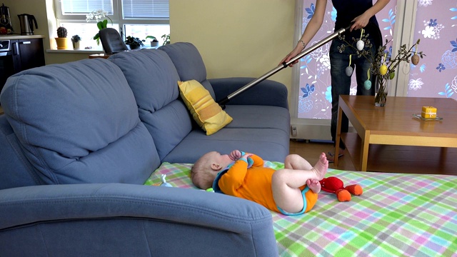 妈妈用吸尘器打扫沙发，小婴儿在旁边玩耍。4 k视频下载