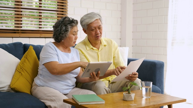 亚洲资深夫妇坐在家里的沙发上用平板电脑搜索关于退休财务文件，资深学会使用科技。就地老化概念视频下载
