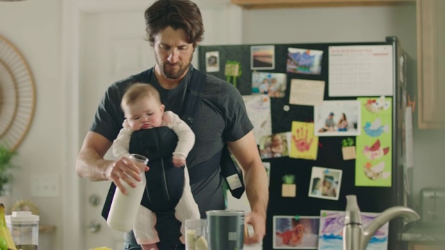 年轻的父亲与孩子在前面面对婴儿背带倒牛奶，同时准备健康的水果奶昔在厨房。视频下载