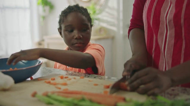 一位非裔美国妇女在厨房柜台上切胡萝卜，儿子帮她把胡萝卜舀进搅拌碗里。视频下载