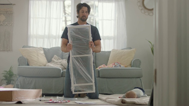 年轻的父亲在组装平板家具，而可爱的婴儿在毯子上滚来滚去。视频素材