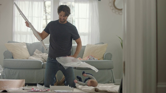 年轻的父亲在客厅组装平板家具，婴儿伸手去抓他。视频素材