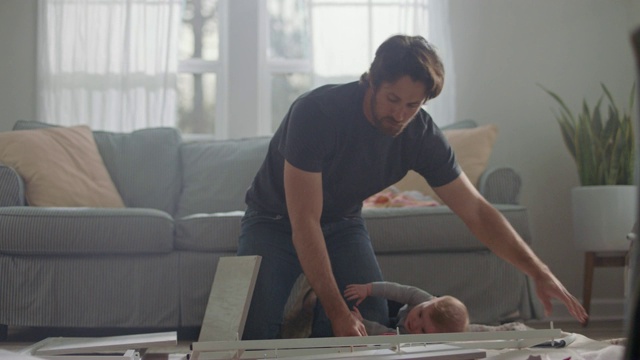金毛猎犬穿过屏风，年轻的父亲试图组装平板家具，婴儿伸手去抓他。视频素材