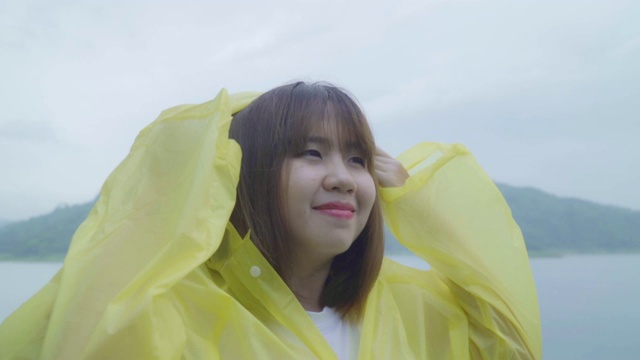 慢动作-年轻的亚洲妇女感到快乐的雨，而穿着雨衣站在湖边。女性在雨天享受和放松的生活方式。视频素材