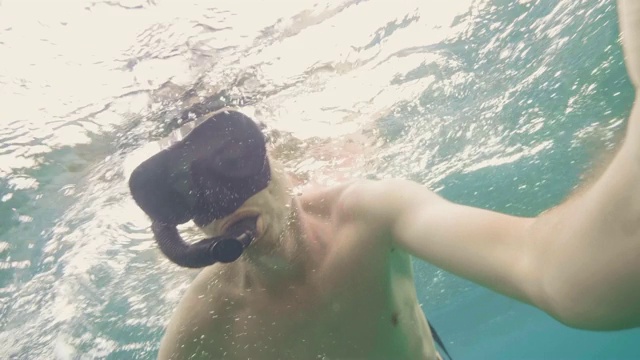 一名年轻男子戴着通气管和面罩在海上浮潜并拍摄自拍视频。一个人在珊瑚礁和鱼的水下游泳，看着镜头。潜水和浮潜。视频素材