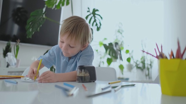 一个穿着蓝色衬衫的微笑男孩坐在客厅的桌子上用铅笔在纸上画画视频素材