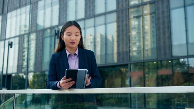 一位年轻的亚洲商务女性在一座现代商务大楼前用平板电脑滚动屏幕视频素材