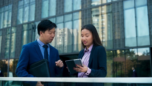 一名亚洲商务女性在办公大楼前一边滚动平板电脑，一边和她的亚洲男同事交谈视频素材