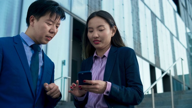 一名亚洲商务女性在办公大楼前向她的男同事展示她手机上的数据，并寻求建议视频素材