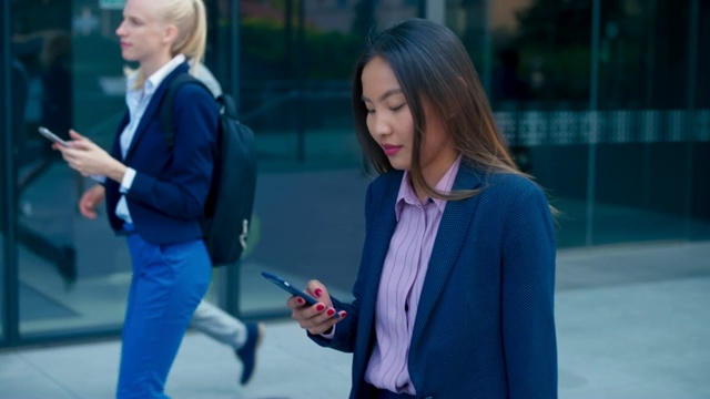 亚洲商务女性在一座现代商务大楼外边走边刷智能手机视频素材