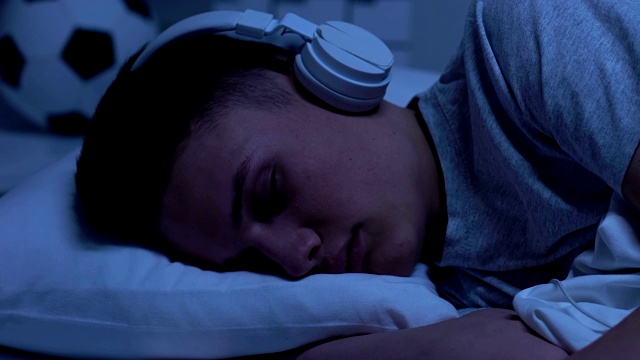 男青少年戴着耳机睡在家里，听有声书，学习视频素材