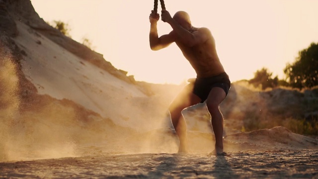 在强大的体能训练中，一个带着带扣的身体膨胀的人用力地击打地面上的绳索，扬起灰尘和阳光视频下载