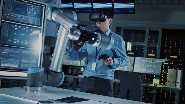 专业的日本开发工程师在蓝色衬衫控制一个未来的机器人手臂与虚拟现实耳机和操纵杆在高科技研究实验室与现代设备。视频下载