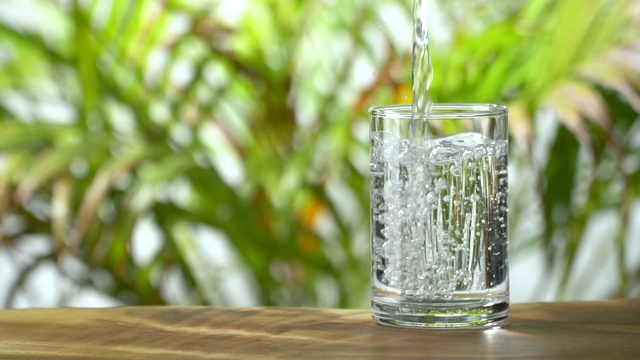 慢动作:把水倒进一个水杯里，背景是茂盛的树叶。视频素材