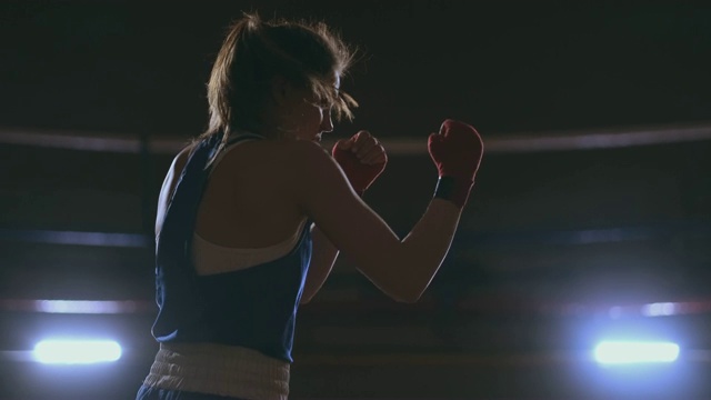 职业美丽的女拳击手在一个黑暗的大厅里，穿着蓝衣服，手腕上缠着红色绷带，用慢镜头进行了一场影子格斗。替身拍摄视频素材