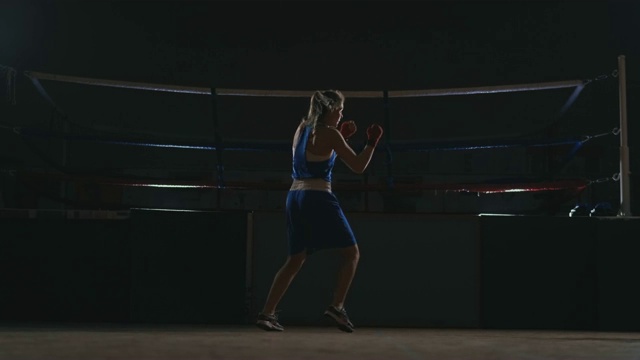 漂亮的职业女拳击手otbryvatyvat穿着蓝色的衣服，手腕上缠着红色的绷带，在大厅的一个黑暗的大厅里，用慢镜头与一个影子搏斗。绕着物体飞行。替身拍摄视频素材