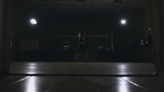 漂亮的职业女拳击手otbryvatyvat穿着蓝色的衣服，手腕上缠着红色的绷带，在大厅的一个黑暗的大厅里，用慢镜头与一个影子搏斗。变焦镜头。替身拍摄视频下载