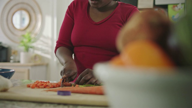 一位非裔美国妇女将胡萝卜切成小块，然后舀进厨房柜台上的搅拌碗里。视频下载