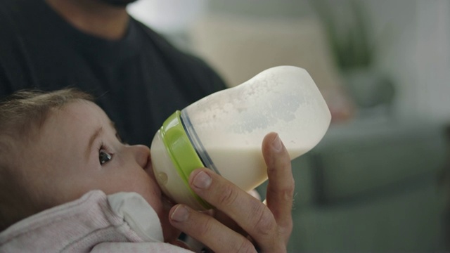 宝贝女儿被父亲抱在怀里，喝着奶瓶里的牛奶。视频素材