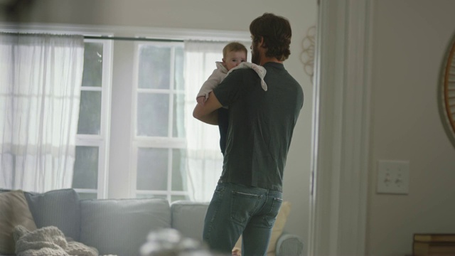 年轻的父亲在阳光明媚的客厅里走来走去，拍拍宝宝的后背，让她在喂完奶后打嗝。视频下载
