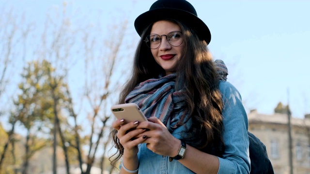 用智能手机给女人发短信。美丽的年轻女子戴着帽子和眼镜使用现代智能手机，在城市中行走，用手机输入短信视频素材