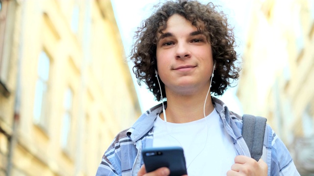 近距离的微笑和大笑英俊卷曲的年轻人听音乐从他的智能手机耳机，走在大街上的老城中心在欧洲视频素材