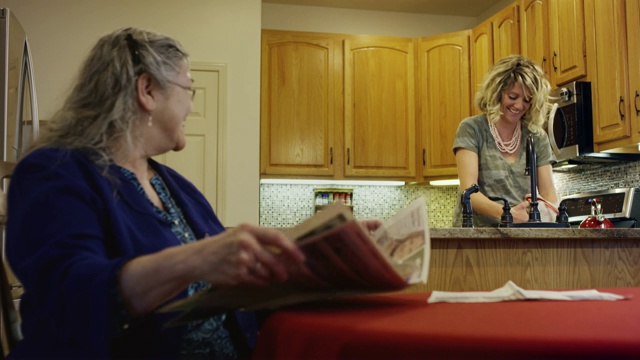 一位70多岁的白人妇女在厨房的桌子上看报纸，她对一位30多岁在水槽里洗碗的白人妇女微笑并交谈视频素材