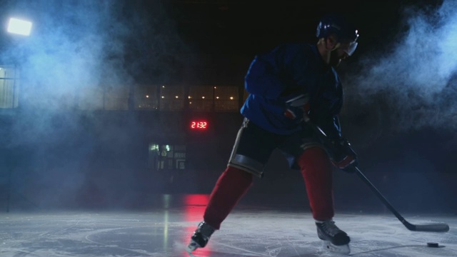 职业冰球运动员拿着一根棒子和一个冰球，在一个黑暗的背景和烟雾中穿着溜冰鞋和头盔在柳达上移动。在冰上场地上用一个年轻人的冰球运球视频素材