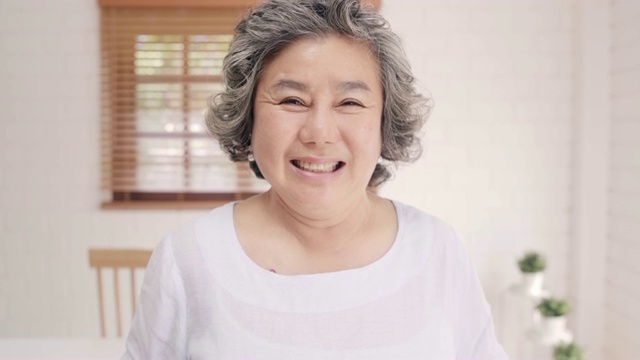 亚洲老年妇女感到幸福的微笑和看着镜头，而放松在客厅的沙发在家里。生活方式高级女性在家的概念。视频下载